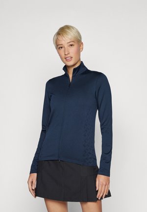Jachetă fermoar bleumarin dintr-un material moale, cu mâneci lungi și un model texturat pe lateral, asortată cu o fustă plisată neagră.