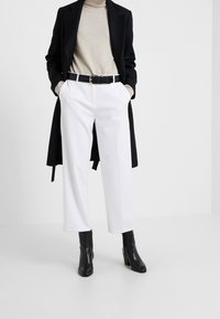 Cappotto nero su dolcevita beige, abbinato a pantaloni bianchi a gamba larga e stivali neri con tacco alla caviglia. Cintura nera con dettagli texture.