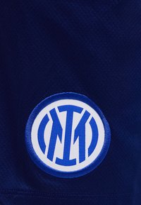Logo brodé circulaire bleu et blanc avec des lettres stylisées entrelacées sur un fond en tissu texturé bleu foncé.