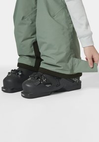 Botas de esquí negras con múltiples hebillas y suelas de goma texturizadas. Monos verde claro con bolsillos laterales y puños ajustables, que se superponen a las botas.