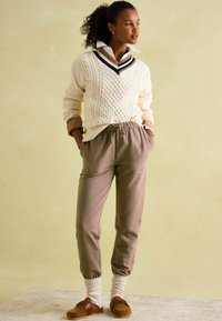 Cremfarbener Zopfmuster-Pullover mit schwarzen und braunen Akzenten, graue Jogginghosen, cremefarbene Socken und braune Slipper, vor einem hellen Hintergrund.