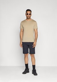 Camisa beige de manga corta, pantalones cortos azul marino, calzado negro y gafas de sol, de pie frente a un fondo blanco liso.