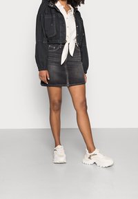Chaqueta negra corta con puños elásticos sobre una camisa blanca atada, combinada con una falda de mezclilla negra. Zapatillas blancas con acentos de estampado animal.