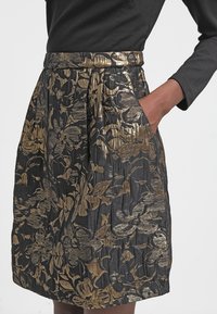 Jupe noire taille haute avec motifs floraux dorés métalliques, plis et poches latérales, portée avec un haut noir à manches longues.