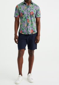 Kortärmad blommig skjorta i lila med gröna blad och röda/gula blommor, i kombination med marinblå shorts och vita sneakers.