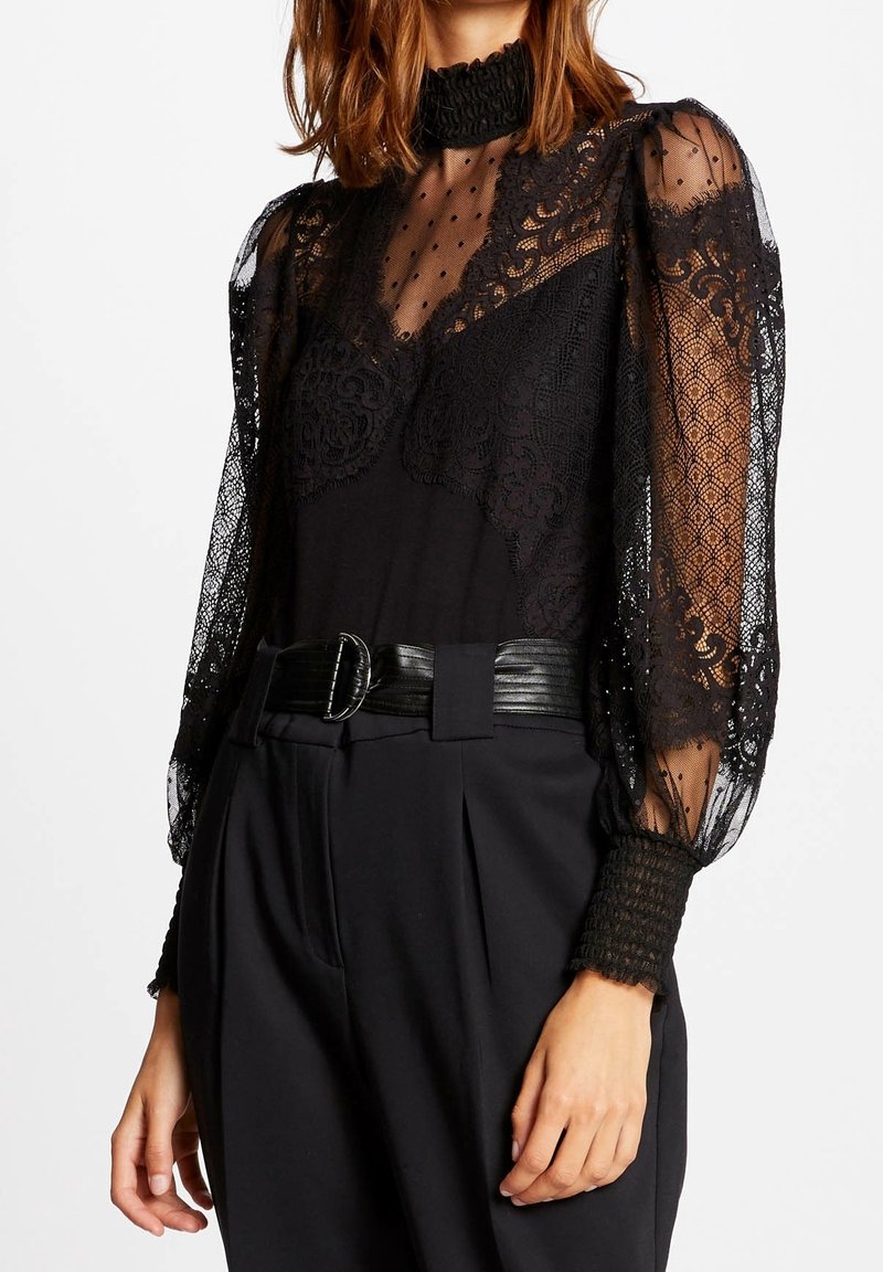 Femme portant un haut en dentelle noir à col montant avec manches bouffantes transparentes et un pantalon noir taille haute avec une large ceinture.