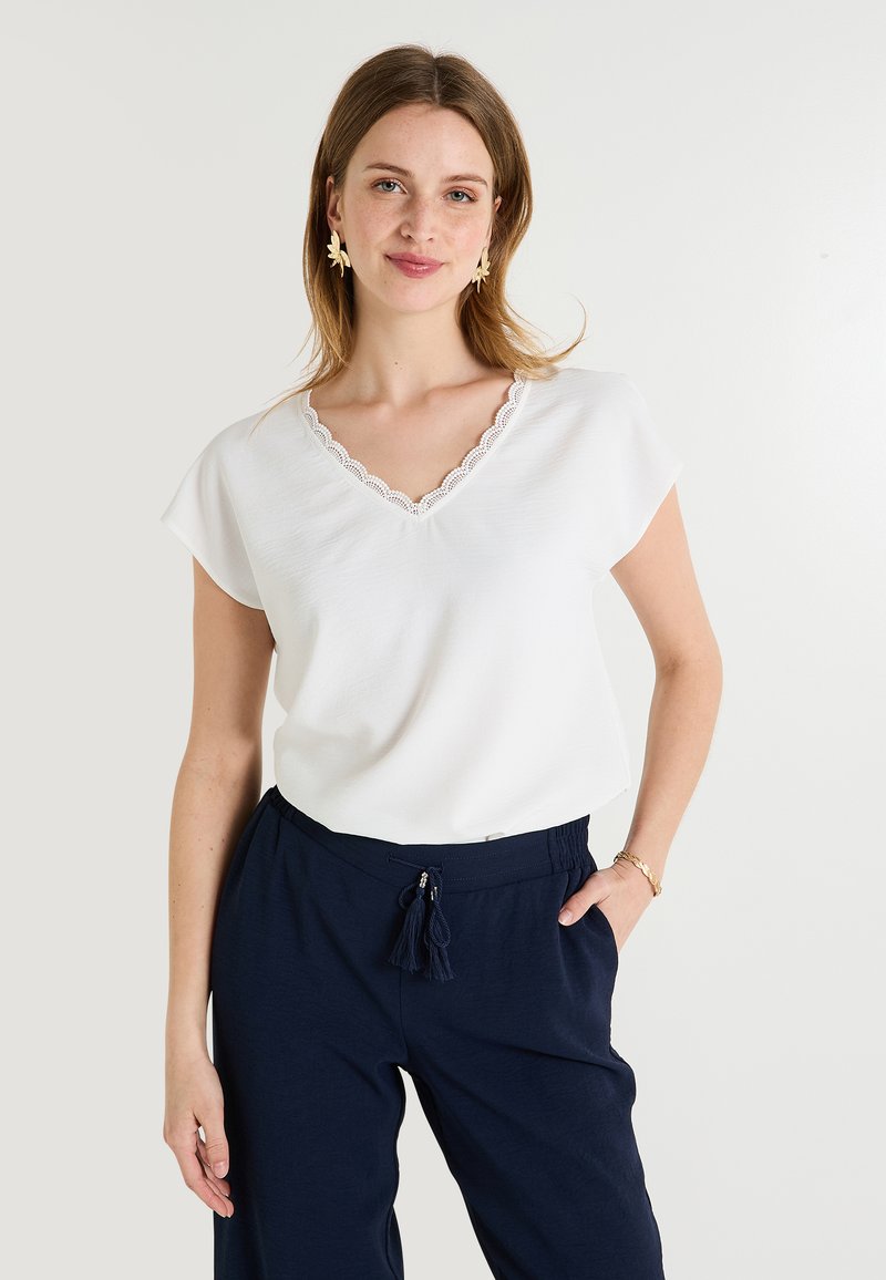 Haut blanc à manches courtes et décolleté en V avec une bordure en dentelle. Associé à un pantalon marine avec cordon de serrage et accents à pompons.