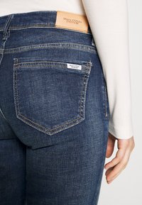 Jean en denim bleu foncé avec une finition texturée, comportant des poches arrière, une étiquette en cuir sur la ceinture et des accents de couture visibles.