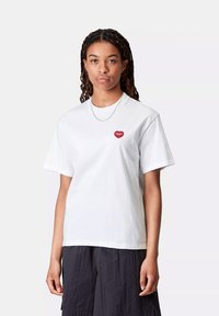 Młoda osoba z warkoczami ma na sobie białą koszulkę z małym czerwonym logo serca i ciemne, teksturowane spodnie, stojąc na jednolitym tle.