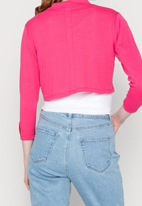 Cropped, long-sleeve shirt in bright pink fabric, with rounded hem. Worn over a white top and paired with light blue, high-waisted jeans.