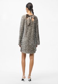 Μίνι φόρεμα με παγιέτες σε ασημί χρώμα, με μακριά μανίκια και βαθύ V-ντεκολτέ στην πλάτη που διαθέτει φιόγκο. Λείο υλικό και ευθεία κοπή στο τελείωμα.