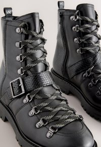 Mustat nahkaiset nilkkurit, joissa on teksturoidut yksityiskohdat, metalliset soljet ja ny öritys edessä. Varustettu sivuvetoketjulla ja karkealla pohjalla.