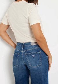 Camiseta ajustada beige de mangas cortas, combinada con jeans de cintura alta de color azul oscuro que presentan un logo triangular de Guess en el bolsillo trasero.