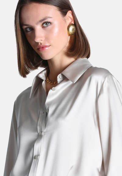 Femme aux cheveux bruns courts portant une chemise blanche brillante boutonnée, de grandes boucles d'oreilles en or et un collier en chaîne dorée, regardant légèrement sur le côté.