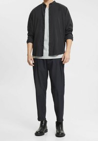 Camicia nera con colletto alto, indossata sopra una camicia grigia chiaro, abbinata a pantaloni scuri a gamba affusolata e stivaletti neri alla caviglia.