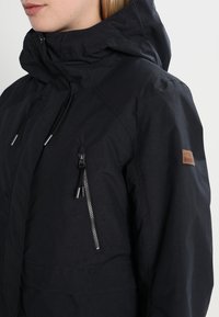 Zwarte waterdichte jas met een hoge kraag, verstelbare capuchon en zijzakken met rits. Beschikt over een tan leren logo-label.