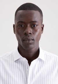 Camicia bianca con sottili righe verticali grigio chiaro, dotata di collo con bottoni e tessuto liscio. Il volto del modello è neutro e ben illuminato.