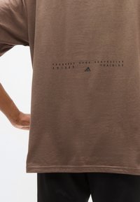 Καφέ βαμβακερό μπλουζάκι με κοντά μανίκια, με μαύρο τύπωμα που γράφει "ΠΡΟΟΔΟΣ ΠΑΝΩ ΑΠΟ ΤΗΝ ΤΕΛΕΙΟΤΗΤΑ" και το λογότυπο της Adidas στην πλάτη.