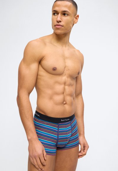 Μυώδης άνδρας όρθιος χωρίς πουκάμισο, φορώντας ριγέ μπλε, πορτοκαλί και μωβ boxers με μαύρη ζώνη μέσης και λευκό λογότυπο σε σκριπτ.