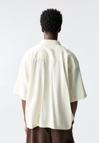 Chemise à manches courtes, boutonnée, de couleur jaune pâle avec de fines rayures verticales. Tissu léger, coupe décontractée et col classique.