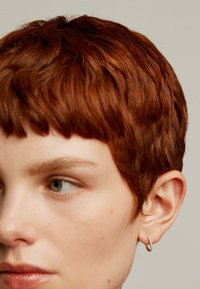 Primer plano de una persona con cabello corto, texturizado y de color castaño rojizo. Arete de aro simple con pequeños adornos en la oreja.