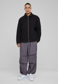 Black zip-up jacket with a collar, paired with loose grey cargo pants. White sneakers complete the outfit. Smooth fabric textures featured.