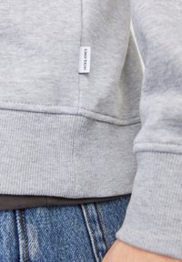 Šedá mikina s žebrovaným lemem a manžetami, nese štítek "JACK & JONES". Nenošena přes tmavou košili a modré džíny z denimu. Měkká textura, ležérní styl.