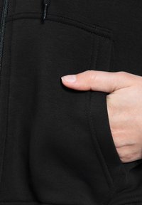 Sweat à capuche zippé noir avec un tissu doux, poche kangourou et poignets côtelés. Main reposant dans la poche, mettant en valeur la texture.
