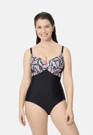 Mujer sonriente con cabello castaño oscuro que lleva un traje de baño de una pieza con parte inferior negra y parte superior con un patrón abstracto y tirantes ajustables.