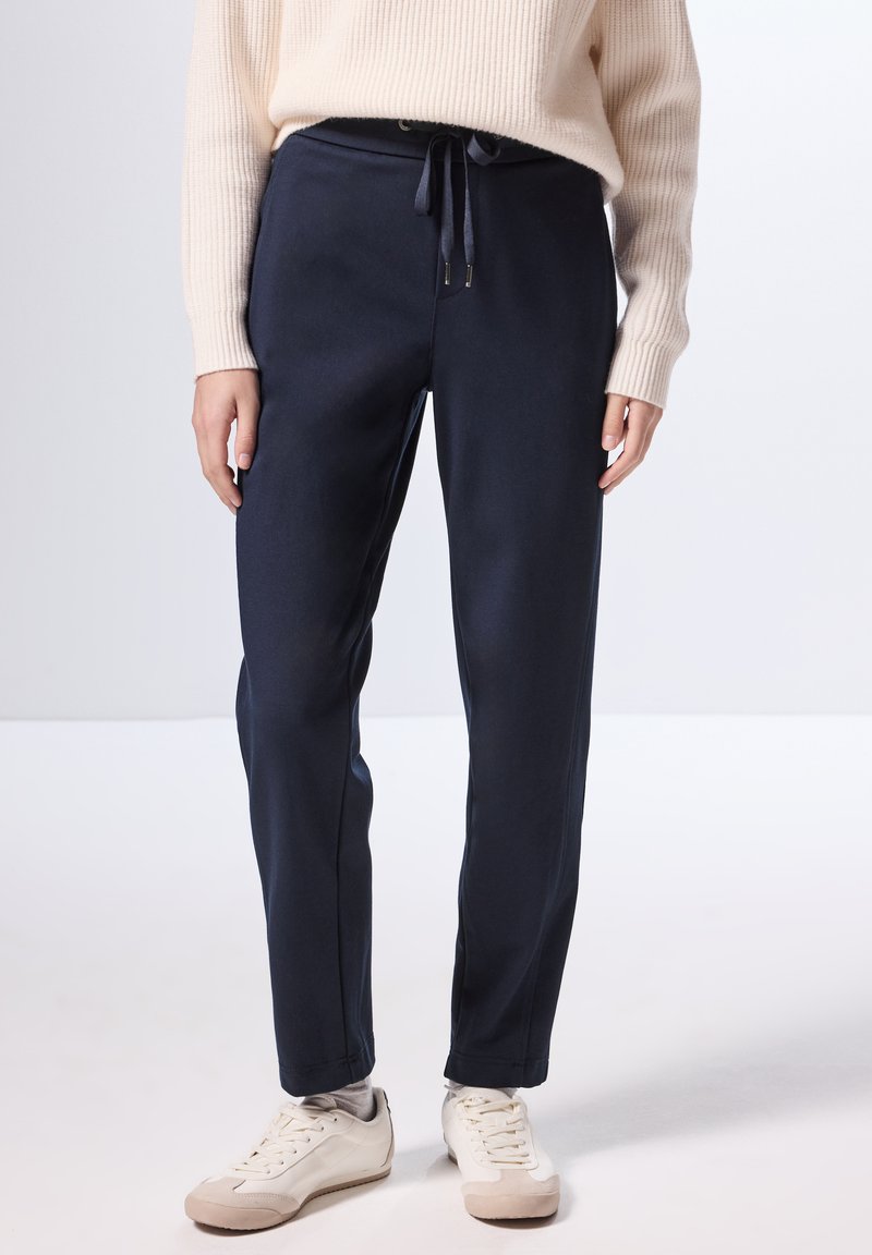 Pantaloni jogger navy in un materiale morbido, con vita regolabile tramite cordoncino, tasche laterali e gambe affusolate, abbinati a sneaker bianche.