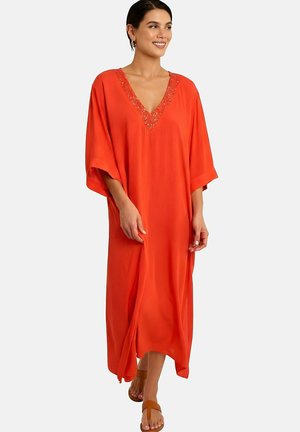 Vrouw in losse, feloranje jurk met versierde V-hals en brede mouwen, lopend op blote voeten met bruine sandalen.