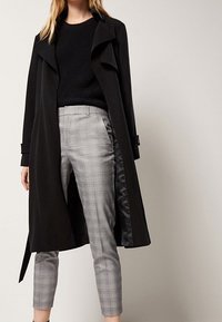 Zwarte trenchcoat met een riem in de taille, grote revers en een satinvoering, gecombineerd met grijs geruite, cropped broek en een gestructureerd zwart sweater.