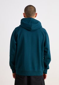 Tyrkysová mikina s jemnou textúrou a klokankovým vreckom. Vybavená veľkou kapucňou a rebrovanými manžetami. Pohľad zozadu zdôrazňuje minimalistický dizajn.