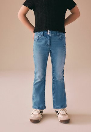 Person iført lyseblå jeans med dobbelte knapper, sort tekstureret kortærmet top og hvide sneakers med sorte striber, stående med hænderne bag ryggen.