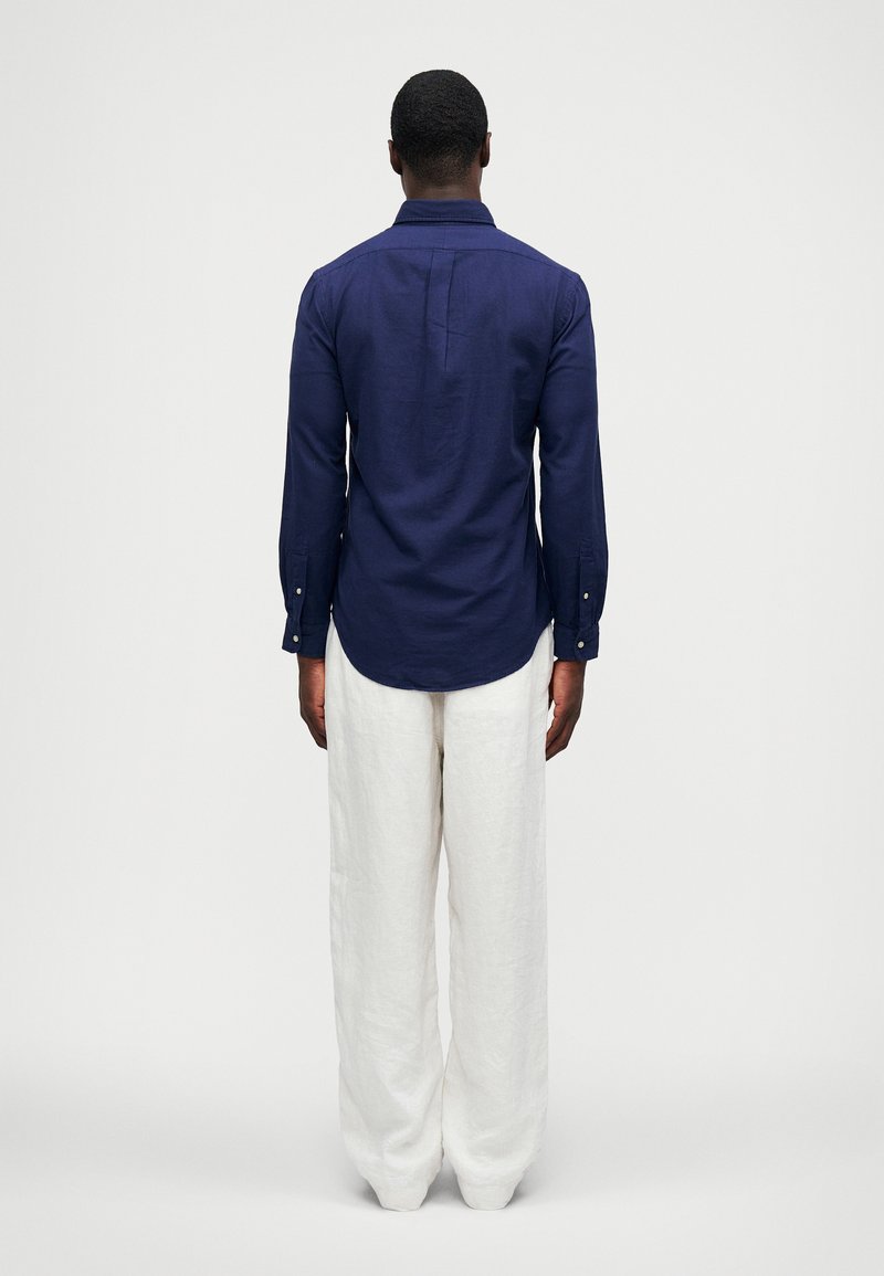 Mørkeblå langærmet skjorte i linned med knapmansjetter, parret med hvide linned bukser med brede ben, set fra ryggen.