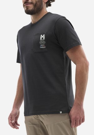 Man wearing black short-sleeve t-shirt with front pocket and logos, paired with beige pants, shown from chest to mid-thigh.