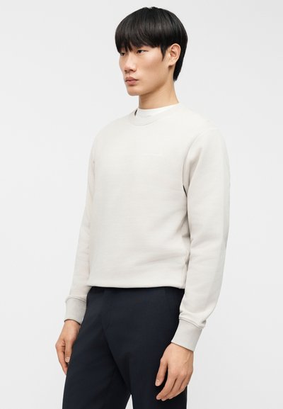 Νεαρός άνδρας που στέκεται, φοράει ανοιχτό μπεζ φούτερ και σκούρο παντελόνι, μπροστά σε απλό λευκό φόντο.