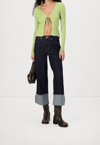 Hellgrünes, geripptes Crop-Top mit Krawattenverschluss, hochgeschnittene, dunkelblaue Jeans mit umgeschlagenen Säumen und braune Stiefeletten mit Schnallen.