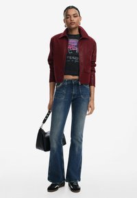 Veste en cuir bordeaux, courte, avec un large col. T-shirt graphique noir, jeans évasés bleus, sac à main noir et chaussures multicolores.