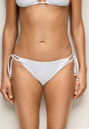 Kobieta w białym, haftowanym bikini z wiązaniami po bokach, stojąca na jednolitym jasnym tle, widoczne środkowa część ciała i górna część nóg.