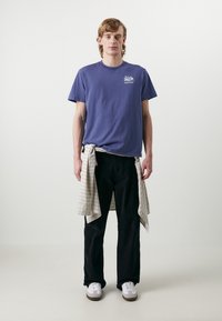 Camiseta de algodón morada con un pequeño gráfico, combinada con pantalones negros. Una camisa a cuadros atada a la cintura y zapatillas blancas.