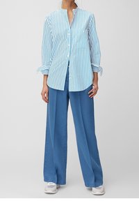 Camicia a righe blu e bianca con colletto e taschino sul petto, abbinata a pantaloni blu a gamba larga. Il modello indossa sneakers bianche.