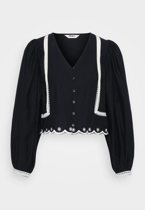 Blouse noire à manches bouffantes et coupe courte, avec une dentelle le long du décolleté et des poches, ornée d'une broderie florale blanche à l'ourlet.