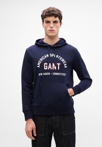 Granatowa bluza z kapturem wykonana z mieszanki bawełny, z przednią kieszenią, kaptur ze sznurkiem i nadrukiem logo w białym i czerwonym kolorze. Proste, ściągacze na rękawach.