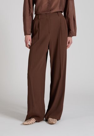 Mujer vistiendo pantalones marrones de talle alto y pierna ancha, combinados con un top marrón de manga larga a juego y zapatos planos tejidos beige.