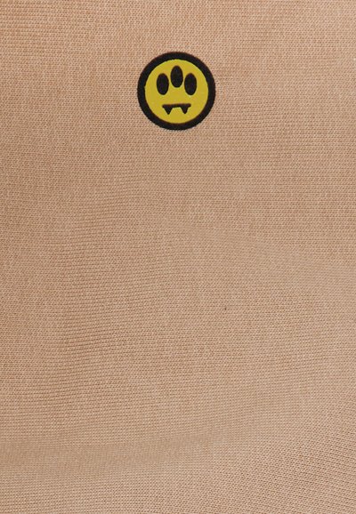 Fondo textil beige con un diseño de cara sonriente amarilla con círculos negros, que presenta tres ojos y colmillos, creando un detalle juguetón y único.