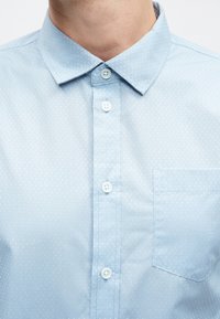 Világoskék, gombos ing apró pöttyös mintával, hegyes gallérral és egyetlen zsebbel a bal oldalon. A gombok fehérek.