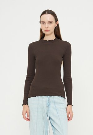 Femme aux longs cheveux bruns portant un haut cintré à manches longues marron foncé avec des bords volantés et un jean bleu clair sur un fond uni.