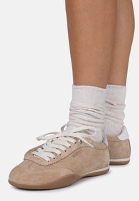 Beige mokkasiner i semsket skinn med hvite lisser og detaljer. Har en teksturert overflate og en gummisåle. Brukt med hvite ribbede sokker.