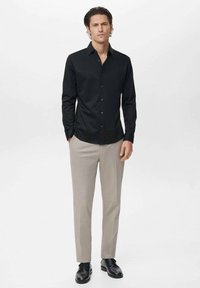 Un hombre está de pie vistiendo una camisa negra abotonada, pantalones beige a medida y zapatos de vestir negros, frente a un fondo blanco liso.